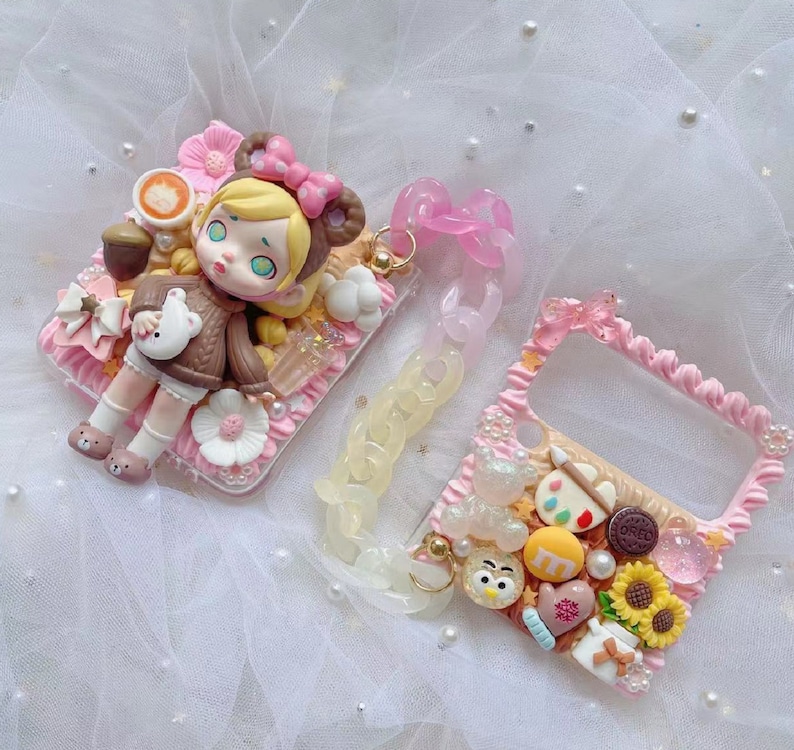 May include: Two decorative phone cases with a whimsical design. One features a doll figure in a brown sweater, the other has a handle and is decorated with sweets and a sunflower. Both cases are pink and white with pearl accents.