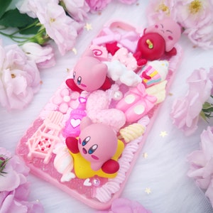以下が含まれることがあります： ピンクの携帯電話ケースは、ピンクと白の3D装飾で飾られています。ピンクのハート、ピンクのバレリーナシューズ、ピンクと白のキャンディーケーン、ピンクの漫画のキャラクター3体が含まれています。ケースはピンクのバラで囲まれています。