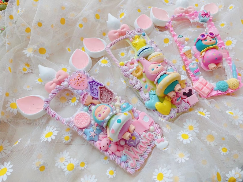 May include: Three decorative phone cases with whimsical designs. Each case features a unique arrangement of colorful, sculpted elements, including bows, stars, and cartoon characters. The cases are in shades of pink, blue, and yellow, with a textured, decorative border.