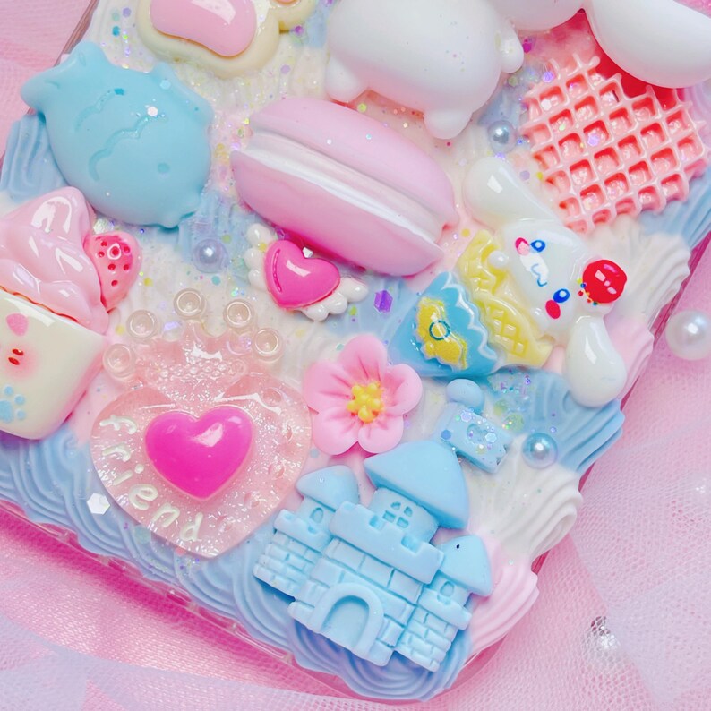 Pu&ograve; includere: Una custodia per telefono rosa e blu decorata con una variet&agrave; di ciondoli, tra cui un castello, un cuore, un fiore, un waffle e un macaron. La custodia presenta anche glitter e un'iscrizione "friend".