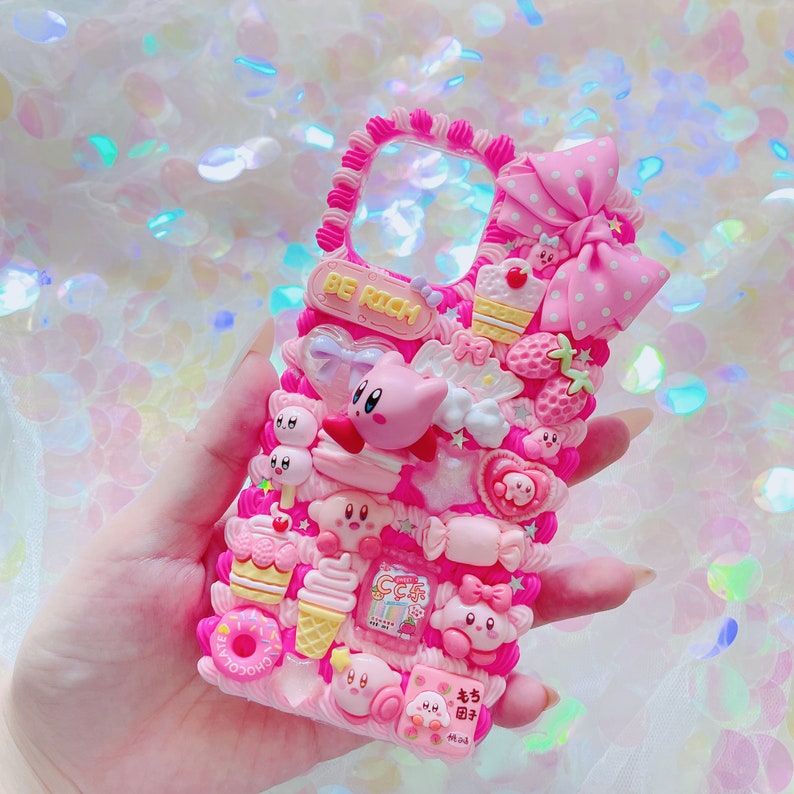 May include: A pink phone case with a raised, textured design. The case is adorned with various 3D embellishments, including a large bow, cartoon characters, sweets, and the text "BE RICH". The overall aesthetic is cute and playful.