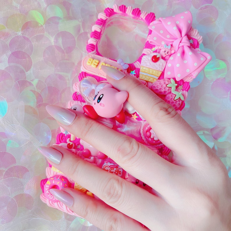 May include: A pink phone case with a decorative design featuring a bow, a cupcake, a strawberry, and a cartoon character. The case has a raised border and is adorned with various embellishments. The text "BE" is visible on a small sign.