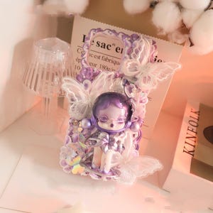 Op de afbeelding: Een decoratieve telefoonhoes met een paarse pop met een ruimtehelm, omringd door kanten vlinders en bloemversieringen. De hoes heeft een lavendelkleurenschema met een papieren achtergrond met de tekst "sac" en "Ce est fabrique".