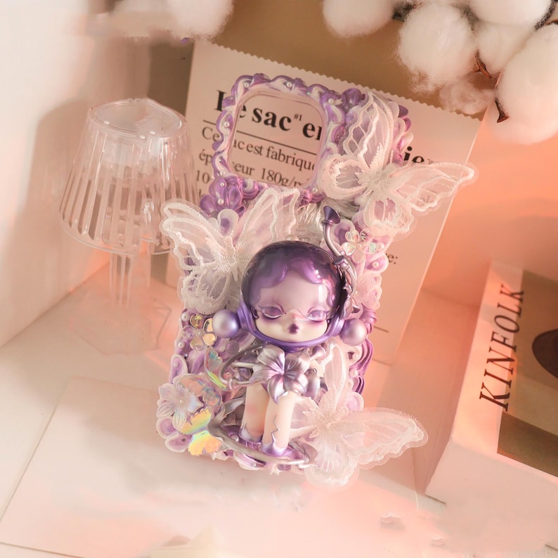 Op de afbeelding: Een decoratieve telefoonhoes met een paarse pop met een ruimtehelm, omringd door kanten vlinders en bloemversieringen. De hoes heeft een lavendelkleurenschema met een papieren achtergrond met de tekst "sac" en "Ce est fabrique".