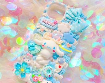 Può includere: Una custodia per telefono bianca e blu decorata con un fiocco blu, una nuvola, una ciambella, una stella, un cuore, un pianeta, un razzo e un piccolo cane bianco con un fiocco blu. La custodia ha anche le parole "Pretty" e "Chocolate" scritte su di essa.