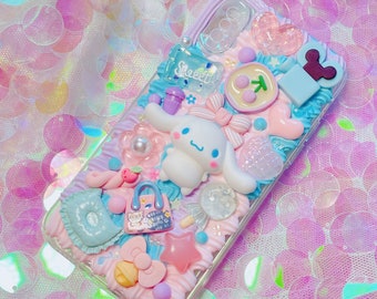 Può includere: Una custodia per telefono trasparente decorata con decorazioni 3D rosa pastello, blu e bianche. La custodia presenta una piccola figurina di cane bianco, una stella rosa, un cuscino blu e una piccola borsa blu con la scritta "Sweet" sopra.