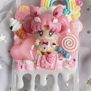 May include: A pink and white phone case decorated with a rainbow, clouds, stars, and a small figurine of Sailor Moon. The case is covered in a white frosting-like substance with pink and white sprinkles.