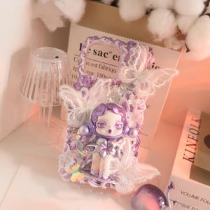 Op de afbeelding: Decoratieve telefoonhoes met een pop met paars haar en een strik, versierd met witte kanten vlinders en paarse versieringen. De hoes heeft een lichtpaars en wit kleurenschema, met een grillig en artistiek ontwerp.