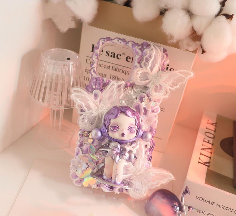 Op de afbeelding: Decoratieve telefoonhoes met een pop met paars haar en een strik, versierd met witte kanten vlinders en paarse versieringen. De hoes heeft een lichtpaars en wit kleurenschema, met een grillig en artistiek ontwerp.