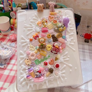 May include: A clear tablet case decorated with colorful, miniature, 3D-style food and character charms. The charms include donuts, ice cream, bears, and other sweets, arranged on a white splash design.