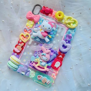 May include: A decorative phone case featuring a pink frame adorned with colorful 3D embellishments. The case includes a pink bow, a Hello Kitty character on a unicorn, and various sweets like macarons and a donut. The background is a soft, white fabric.