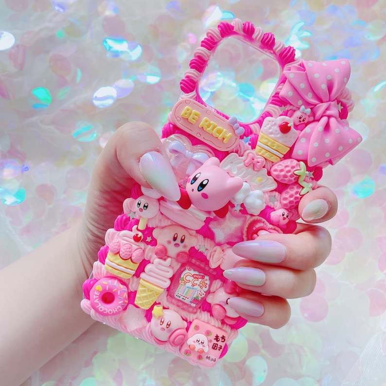 May include: A pink phone case decorated with various charms, including a pink bow, a Kirby character, ice cream, and strawberries. The case has the words "BE RICH" on it.