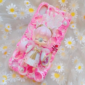 May include: Pink phone case with a 3D design featuring a doll-like figure, butterflies, flowers, and pearls. The case has a textured, swirled background and is decorated with various embellishments. The doll is wearing a white and gold outfit and holding a musical instrument.