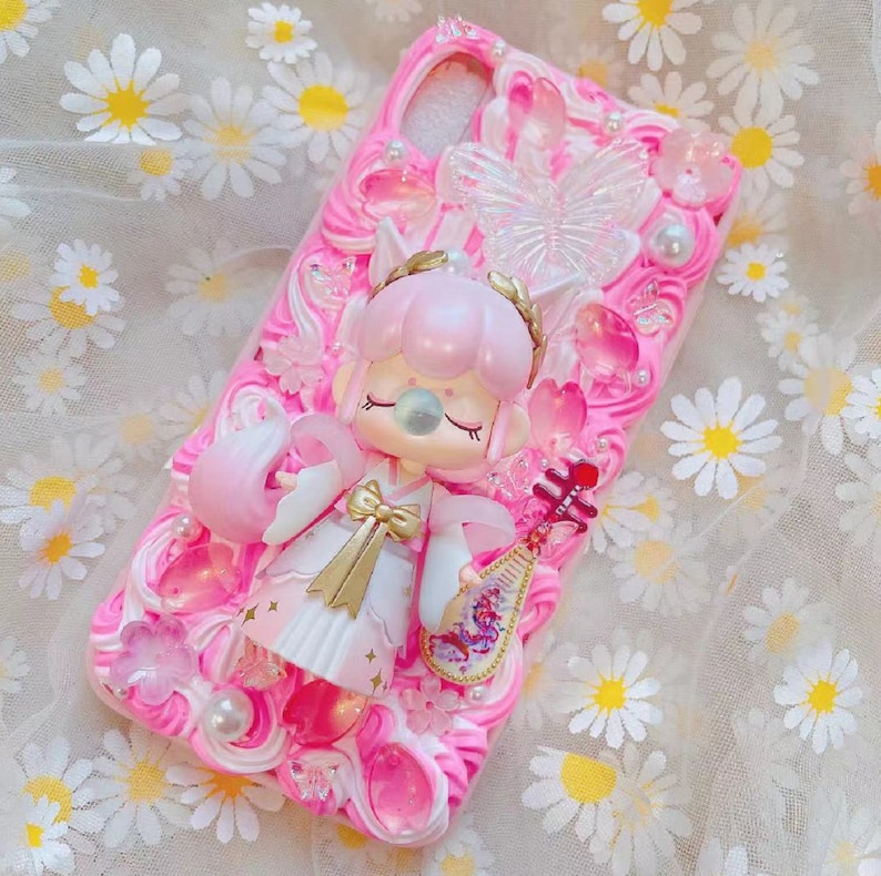 May include: Pink phone case with a 3D design featuring a doll-like figure, butterflies, flowers, and pearls. The case has a textured, swirled background and is decorated with various embellishments. The doll is wearing a white and gold outfit and holding a musical instrument.