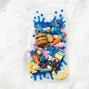 Op de afbeelding: Een transparante telefoonhoes met een blauw, wit en geel ontwerp. De hoes is versierd met verschillende 3D-charms, waaronder een blauw cartoonfiguur, een hamburger, ijshoorntjes en een wafel.