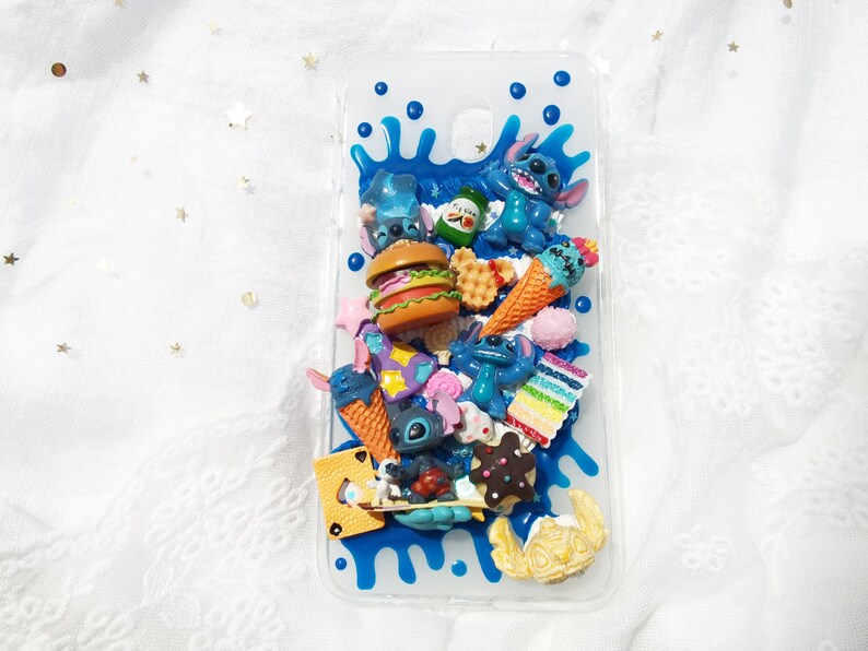 Op de afbeelding: Een transparante telefoonhoes met een blauw, wit en geel ontwerp. De hoes is versierd met verschillende 3D-charms, waaronder een blauw cartoonfiguur, een hamburger, ijshoorntjes en een wafel.