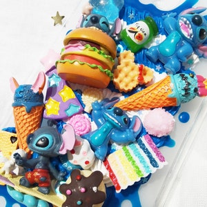 Op de afbeelding: Een blauwe en witte telefoonhoes versierd met verschillende kleurrijke bedels, waaronder een blauw Stitch-personage, een hamburger, een wafel, een ijshoorn en een regenboogtaart. De hoes is gemaakt van helder plastic en heeft een blauw en wit swirl-ontwerp.