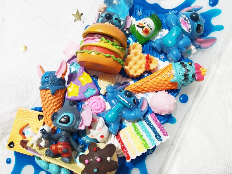 Op de afbeelding: Een blauwe en witte telefoonhoes versierd met verschillende kleurrijke bedels, waaronder een blauw Stitch-personage, een hamburger, een wafel, een ijshoorn en een regenboogtaart. De hoes is gemaakt van helder plastic en heeft een blauw en wit swirl-ontwerp.