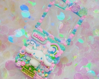 Può includere: Una custodia per telefono trasparente decorata con glassa rosa pastello, blu e verde. La custodia presenta un arcobaleno, nuvole e un cane di cartone animato bianco con la parola "LOVELY" su uno stendardo verde.