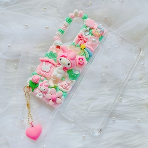 Może przedstawiać: Przezroczyste etui na telefon z różowo-zielonym wzorem przedstawiającym postać Hello Kitty, kwiat i mały domek z napisem "Rabbit". Etui ma złoty łańcuszek z różowym serduszkiem.