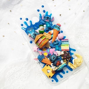Op de afbeelding: Een transparante telefoonhoes met een blauw, wit en geel ontwerp. De hoes is versierd met verschillende 3D-charms, waaronder een blauw personage, ijshoorntjes, een hamburger en een taart.
