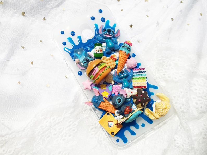 Op de afbeelding: Een transparante telefoonhoes met een blauw, wit en geel ontwerp. De hoes is versierd met verschillende 3D-charms, waaronder een blauw personage, ijshoorntjes, een hamburger en een taart.