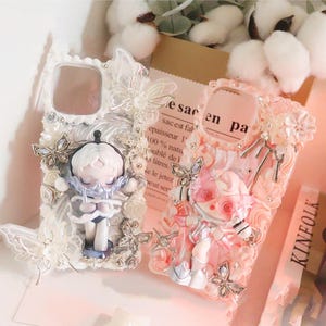 Op de afbeelding: Twee decoratieve telefoonhoesjes. E&eacute;n is wit met een poppenfiguur en vlinderaccenten. De andere is roze met een poppenfiguur en bloemaccenten. De hoesjes hebben een uniek, artistiek ontwerp.