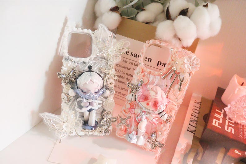 Op de afbeelding: Twee decoratieve telefoonhoesjes. E&eacute;n is wit met een poppenfiguur en vlinderaccenten. De andere is roze met een poppenfiguur en bloemaccenten. De hoesjes hebben een uniek, artistiek ontwerp.