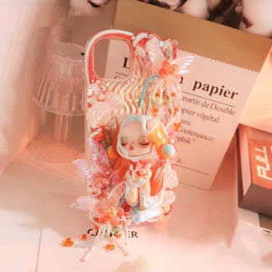 Op de afbeelding: Decoratieve telefoonhoes met een grillig ontwerp. Het heeft een figuur in oranje en wit, versierd met roze en oranje accenten. Het woord 'papier' is zichtbaar op een papieren zak. Een uniek accessoire.