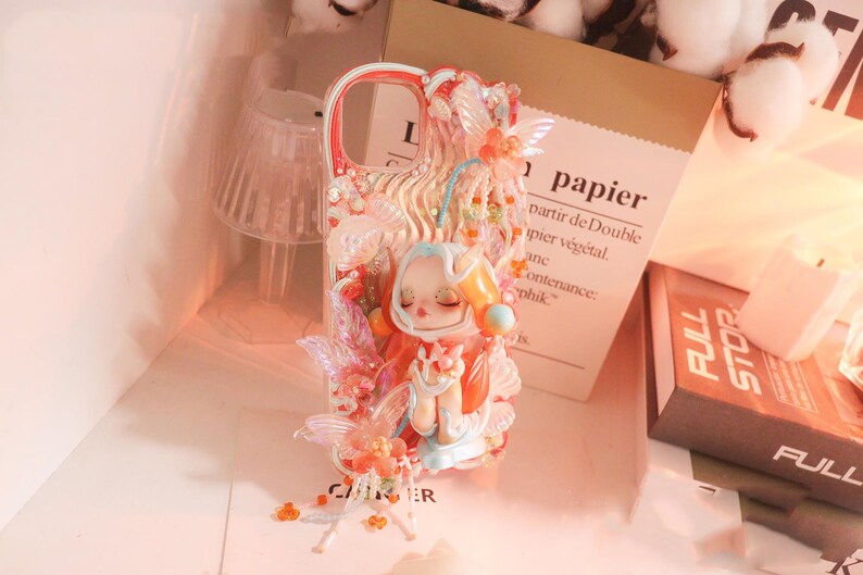 Op de afbeelding: Decoratieve telefoonhoes met een grillig ontwerp. Het heeft een figuur in oranje en wit, versierd met roze en oranje accenten. Het woord 'papier' is zichtbaar op een papieren zak. Een uniek accessoire.