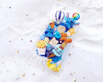 May include: A clear phone case decorated with blue, yellow, and white 3D embellishments. The case features a cartoon character, a hot air balloon, popcorn, a donut, and other fun details.