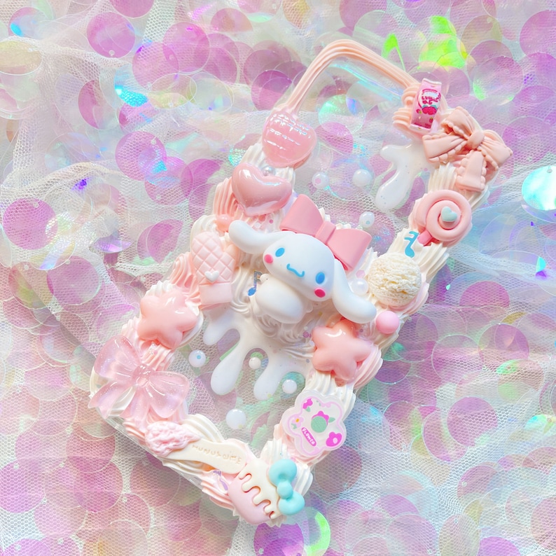May include: A clear phone case decorated with pink, white, and blue 3D embellishments. The case features a white cartoon dog with a pink bow, a pink star, a pink swirl, and a pink lollipop. The case is on a pink and white background.