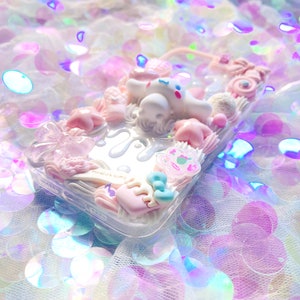 May include: A clear phone case decorated with pink, white, and blue 3D embellishments. The case features a white cartoon character, a pink bow, and a pink star. The text "Harajuku Dime" is visible on the case.