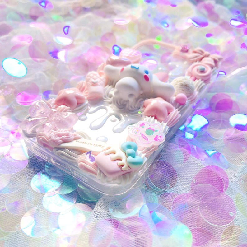 May include: A clear phone case decorated with pink, white, and blue 3D embellishments. The case features a white cartoon character, a pink bow, and a pink star. The text "Harajuku Dime" is visible on the case.