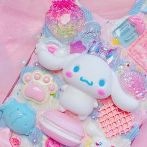 Pu&ograve; includere: Una custodia per telefono rosa e blu decorata con un cane bianco dei cartoni animati, un macaron rosa, un pesce blu, un'impronta di zampa rosa e un waffle rosa e bianco.