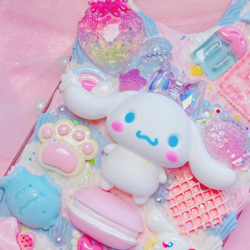 Pu&ograve; includere: Una custodia per telefono rosa e blu decorata con un cane bianco dei cartoni animati, un macaron rosa, un pesce blu, un'impronta di zampa rosa e un waffle rosa e bianco.
