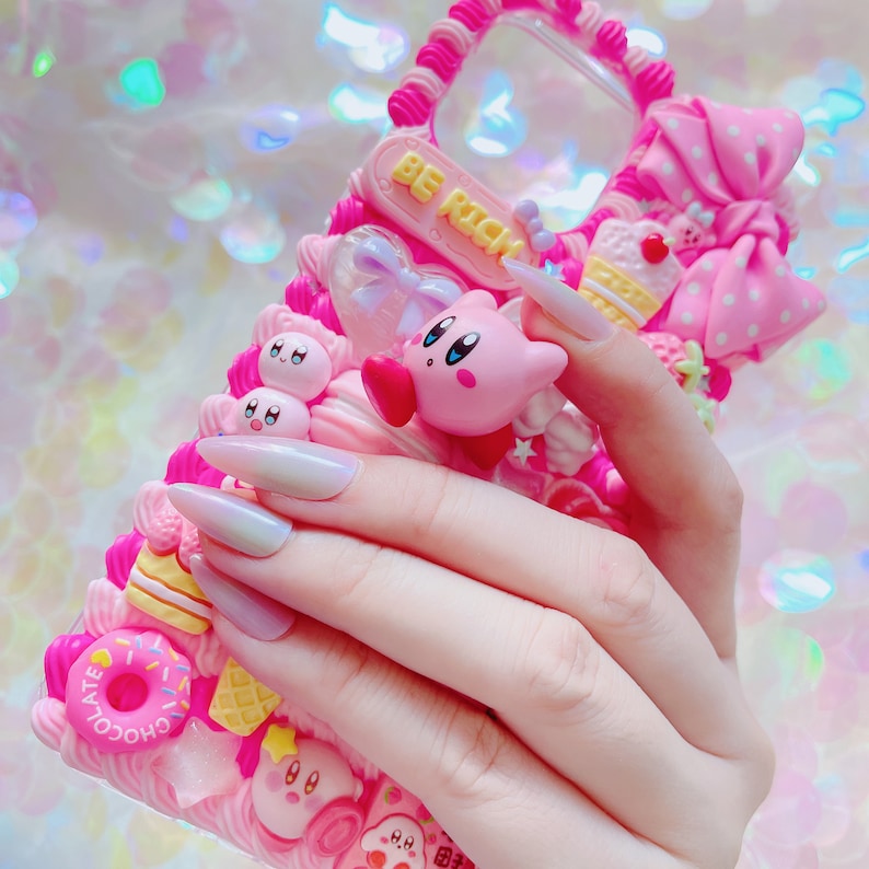 May include: A pink phone case decorated with various 3D elements, including a pink Kirby character, donuts, cupcakes, and a bow. The case has a textured, layered design with the words "BE RICH" on a pink rectangular plaque.