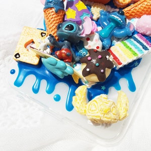 Op de afbeelding: Een blauwe, witte en gele telefoonhoes met een 3D-ontwerp met een blauw Stitch-personage, ijshoorntjes, een regenboogtaart en andere kleurrijke details.