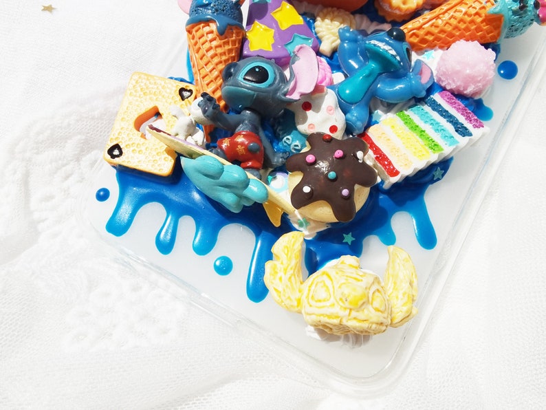 Op de afbeelding: Een blauwe, witte en gele telefoonhoes met een 3D-ontwerp met een blauw Stitch-personage, ijshoorntjes, een regenboogtaart en andere kleurrijke details.