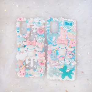 Op de afbeelding: Twee doorzichtige telefoonhoesjes versierd met pastelkleurige versieringen. Elke hoes heeft een wit stripfiguur met grote oren, strikken en diverse bedels, waaronder harten, sterren en desserts. De hoesjes hebben een grillige, handgemaakte uitstraling.