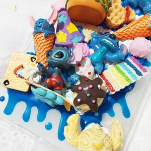 Op de afbeelding: Een blauwe en witte telefoonhoes met een verscheidenheid aan kleurrijke 3D-decoraties, waaronder een blauw Stitch-personage, een bruin en wit koekje en een gele krab.