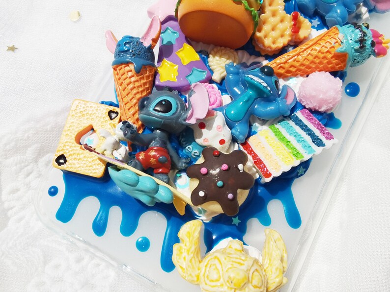 Op de afbeelding: Een blauwe en witte telefoonhoes met een verscheidenheid aan kleurrijke 3D-decoraties, waaronder een blauw Stitch-personage, een bruin en wit koekje en een gele krab.
