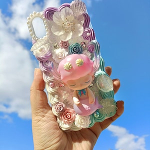 May include: A decorative phone case featuring a sleeping mermaid figurine with a pink hat and tail. The case is adorned with white, pink, and purple floral designs, pearls, and seashells. The case is held against a blue sky background.