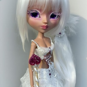 May include: A fashion doll with white hair and purple eye makeup, wearing a white dress with feather accents and a heart-shaped scepter. The doll's dress has a corset-like bodice and a layered skirt. The scepter is gold with a red heart.