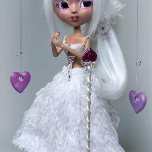 Könnte beinhalten: Eine Puppe mit weißen Haaren und einem weißen Kleid. Die Puppe hat lila Augen und hält ein herzförmiges Zepter. Zwei herzförmige Ornamente in Lila hängen an Schnüren. Das Kleid besteht aus einem flauschigen, strukturierten Material.