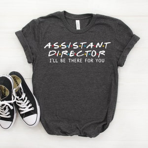 May include: A dark heather grey t-shirt with the text "ASSISTANT DIRECTOR I'LL BE THERE FOR YOU" in a white font. The text is styled to look like the Friends logo.