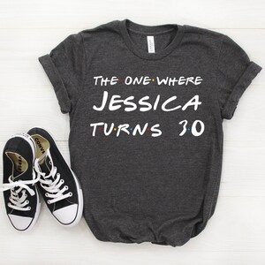 May include: Dark gray t-shirt with the text "THE ONE WHERE JESSICA TURNS 30" in white, with colorful accents. Black canvas sneakers with white laces are next to the shirt. The shirt is folded at the sleeves.