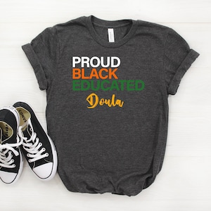 May include: A dark gray t-shirt with the words "PROUD BLACK EDUCATED Doula" printed in white, orange, and green letters.