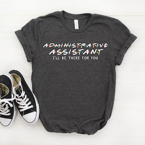 May include: Dark gray t-shirt with the text "ADMINISTRATIVE ASSISTANT I'LL BE THERE FOR YOU" in a white font with colorful dots. The text is styled to look like the title card from the TV show "Friends".