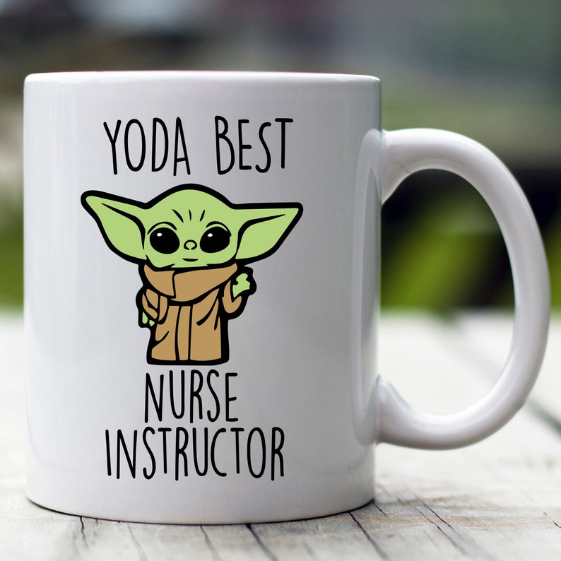 Puede incluir: Taza de cer&aacute;mica blanca con una ilustraci&oacute;n de un personaje de dibujos animados verde y el texto "YODA BEST NURSE INSTRUCTOR" en negro. El personaje tiene orejas grandes y lleva una t&uacute;nica marr&oacute;n. La taza tiene un asa curva.